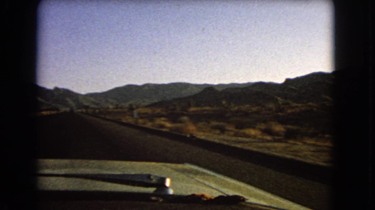 1966 Nevada road-travel frame with the car hood and wiper in front, desert vegetation along the road, and rugged ridges beyond.