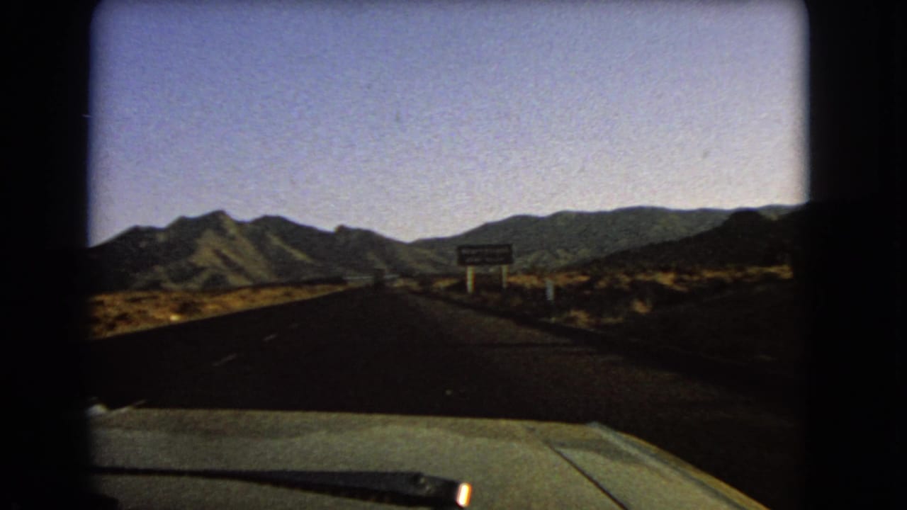 Car hood and desert highway approaching mountains with a roadside sign ahead.