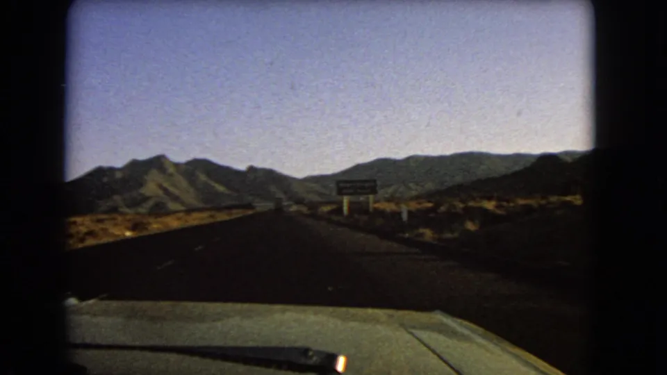 1966 8mm Nevada road view from inside a moving car, with desert hills and a Searchlight exit sign in the distance.