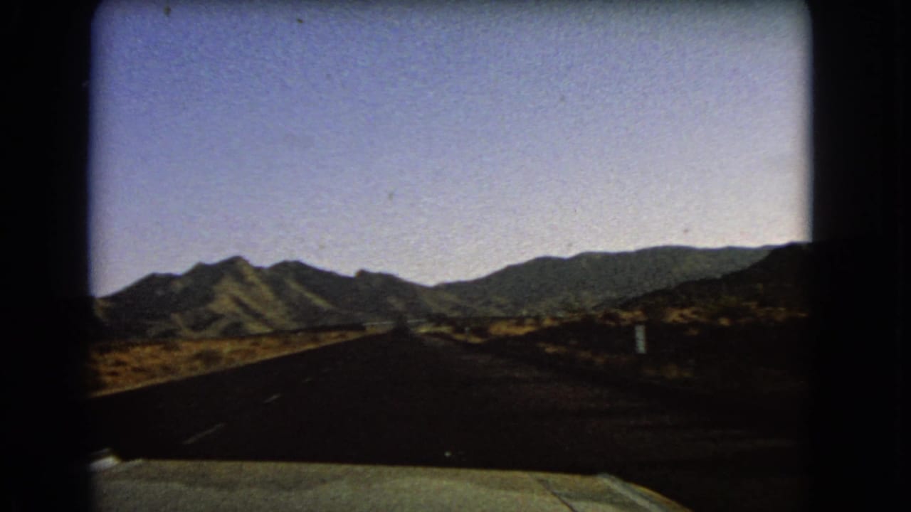1966 Nevada 8mm view from a moving car on a desert road toward low mountain ridges under a pale blue sky.