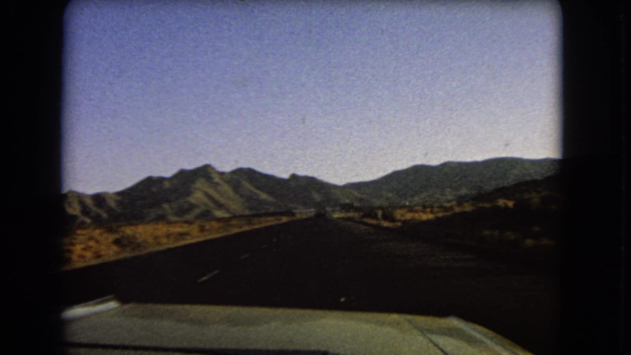 Forward road view through dry terrain with mountain ridges across the horizon.
