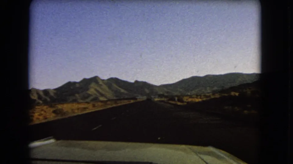 A 1966 Nevada home-movie frame shows a paved road through arid terrain, with dark mountains ahead and the car hood at the bottom edge.