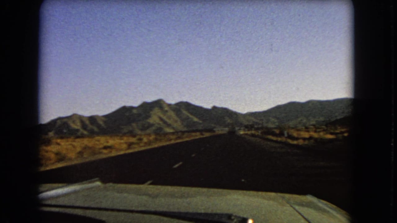 The moving 8mm viewpoint follows a Nevada desert road with arid ground, mountain slopes, and a visible car hood in the foreground.