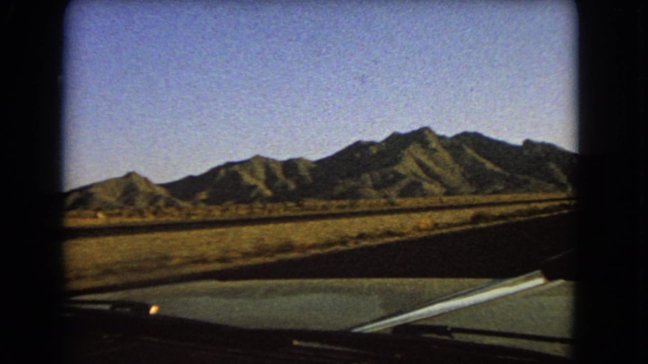 A 1966 Nevada 8mm frame looks from the car toward sunlit desert mountains, dry roadside ground, and a dark paved road edge.