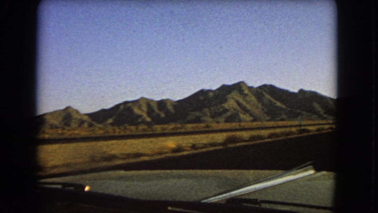 1966 Nevada 8mm windshield view showing a desert road, car hood edge, dry field, and sunlit mountains under blue sky.