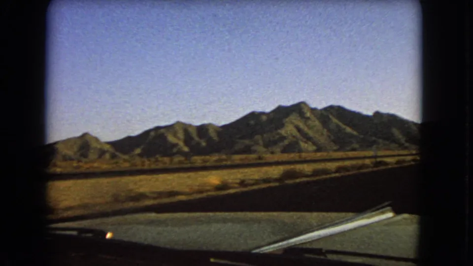 1966 Nevada 8mm windshield view showing a desert road, car hood edge, dry field, and sunlit mountains under blue sky.