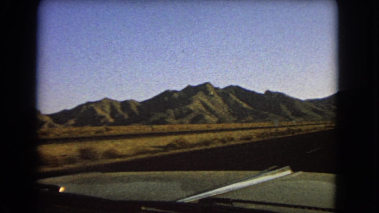 Windshield-framed 1966 Nevada landscape with a paved roadway, dry grasses, and distant mountains filmed from a moving vehicle.