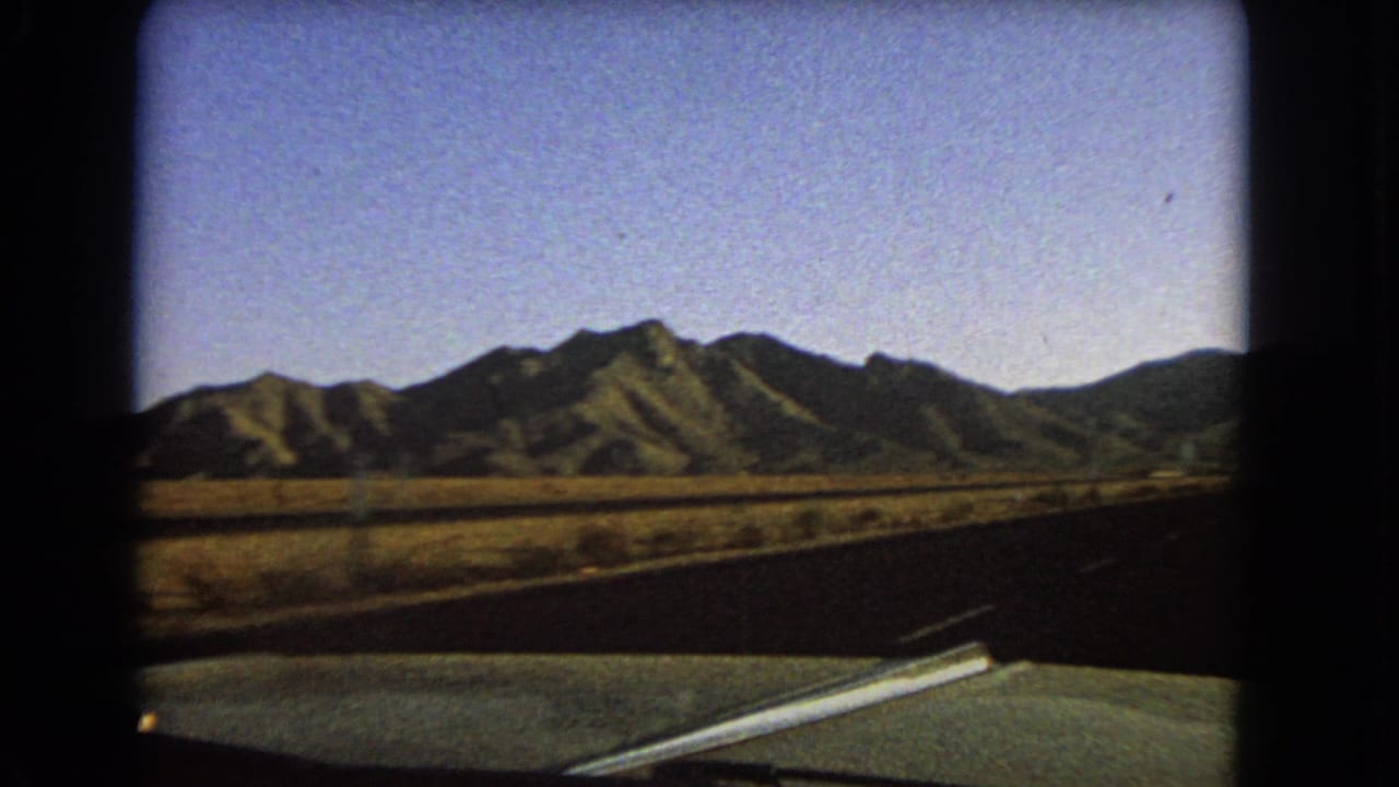 1966 Nevada 8mm travel frame showing a car hood, dark road edge, dry open land, and mountains from a moving vehicle.