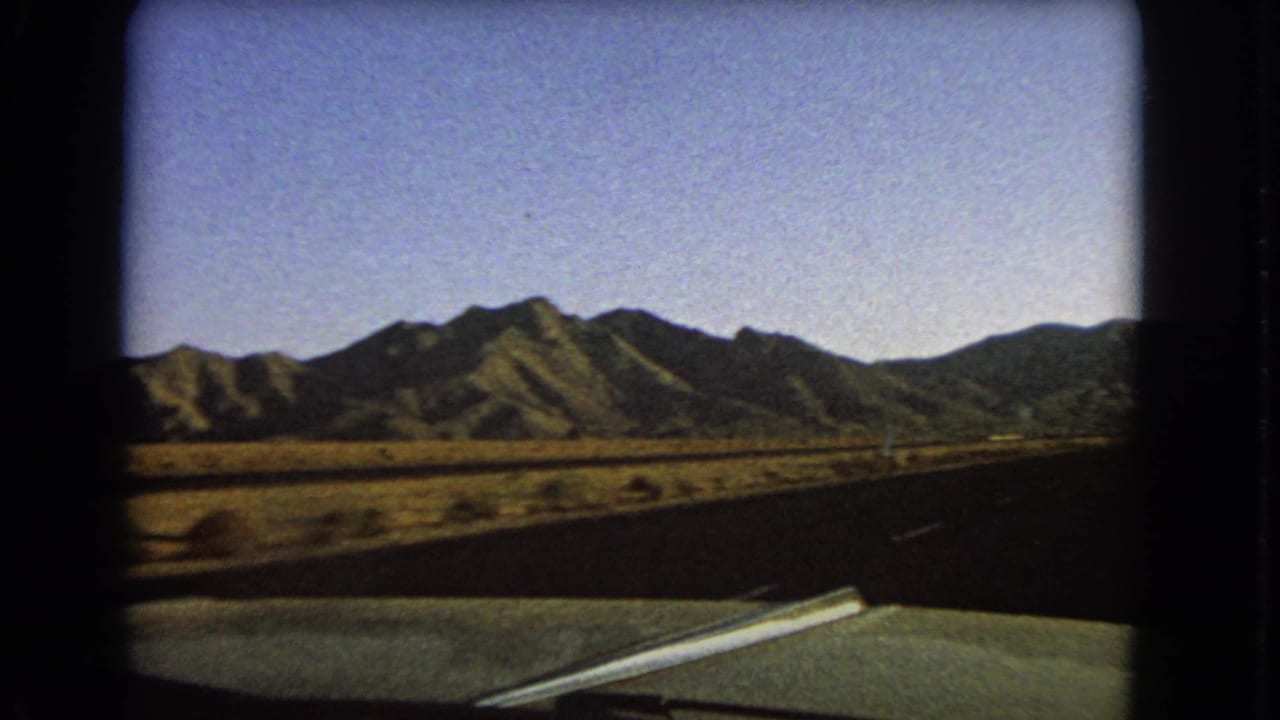 Green car hood and open road facing distant mountains across dry scrubland.