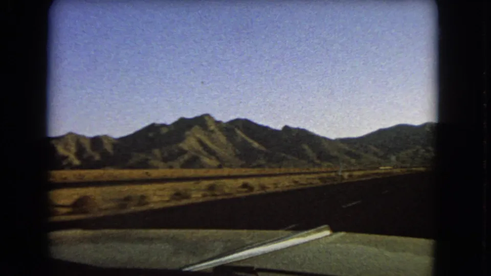 1966 Nevada 8mm frame from inside a green car, looking down an open road toward distant mountains and dry roadside scrub.