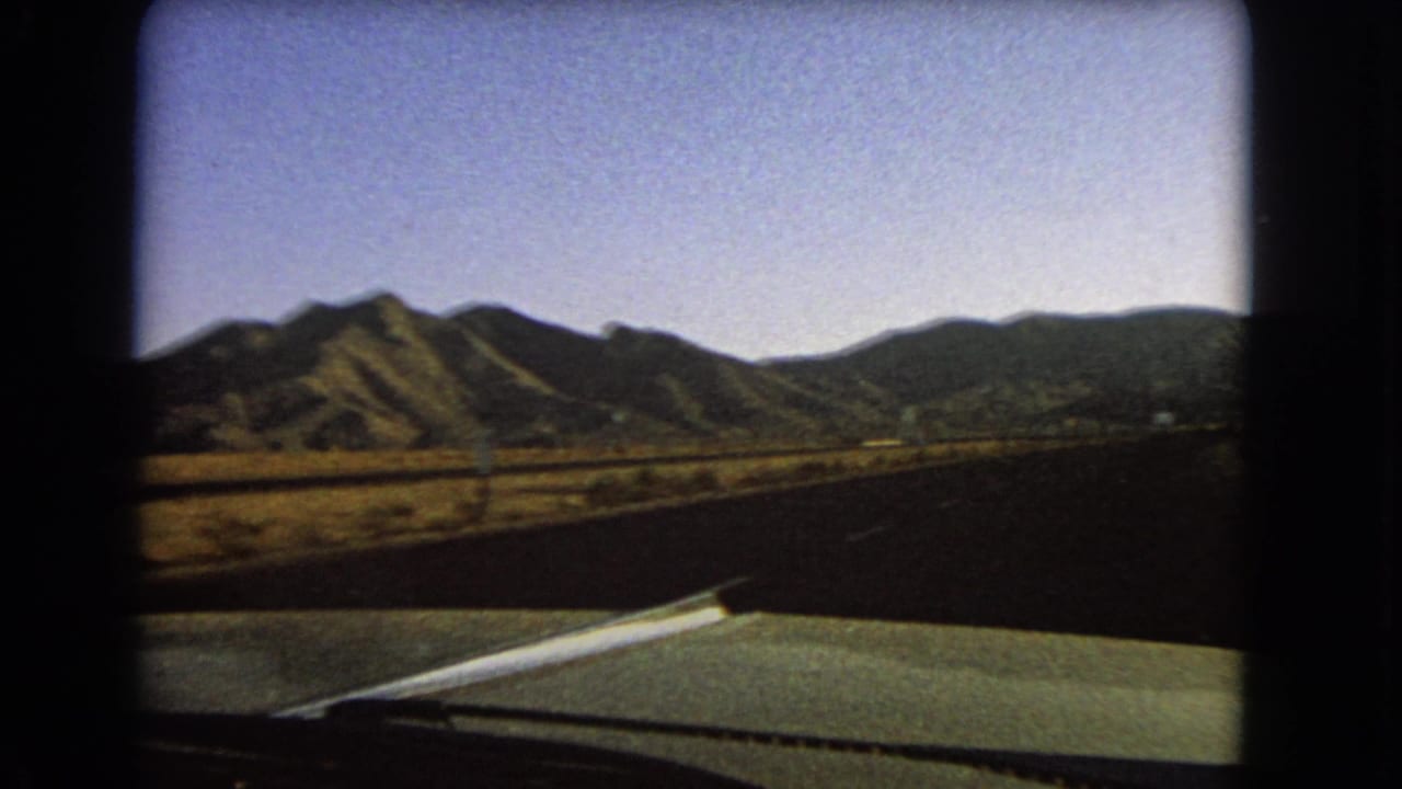 1966 Nevada home-movie view over a green car hood as the road runs through flat desert toward a dark mountain ridge.