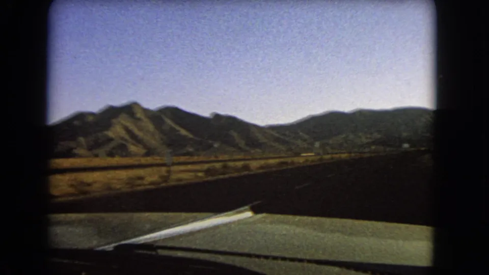 1966 Nevada home-movie view over a green car hood as the road runs through flat desert toward a dark mountain ridge.