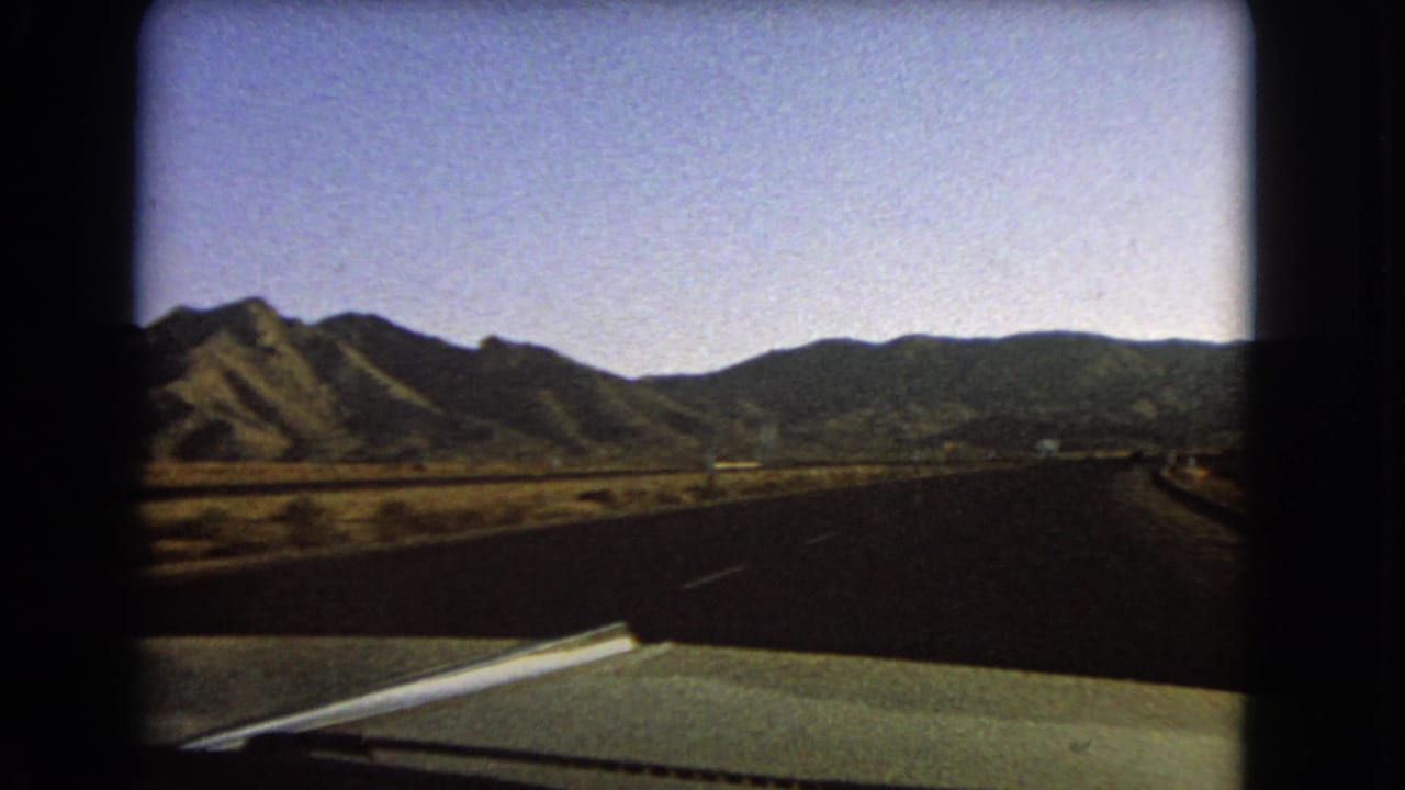 Moving 8mm view from a green car in Nevada, with the highway and shoulder crossing dry open land below distant mountains.