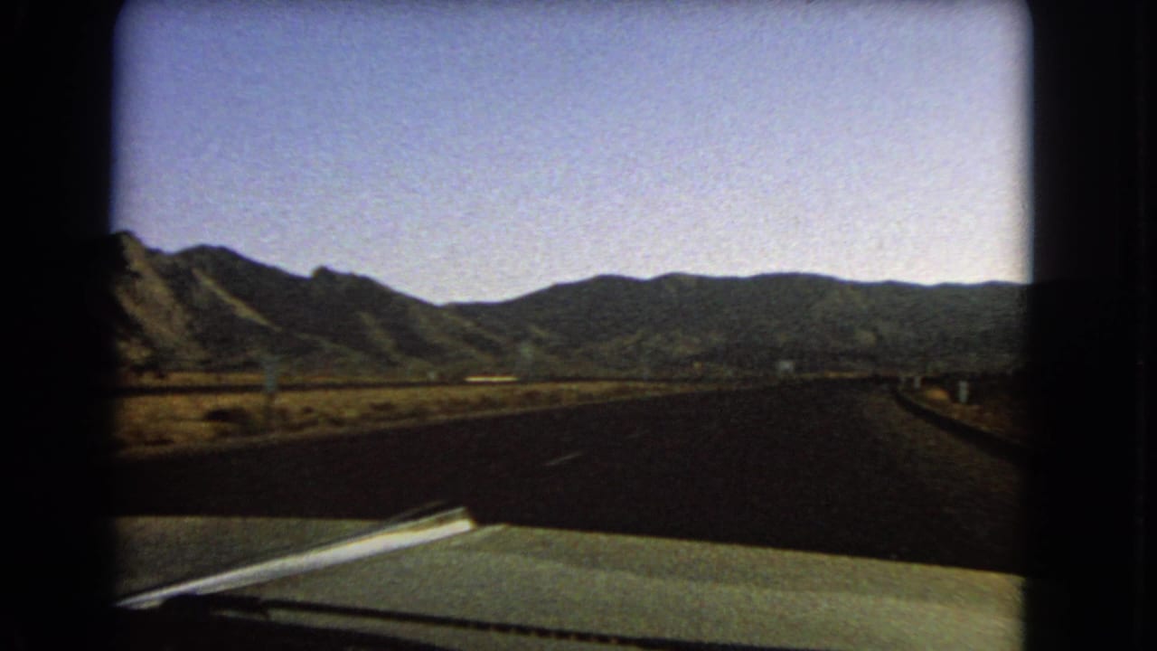 Late-frame 1966 Nevada road view from inside a green car, showing open pavement, sparse scrub and a mountain range ahead.