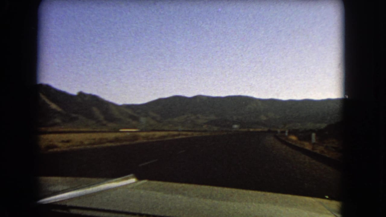 1966 Nevada home-movie frame showing a straight desert road from a car, with distant mountains and the hood or dashboard visible.