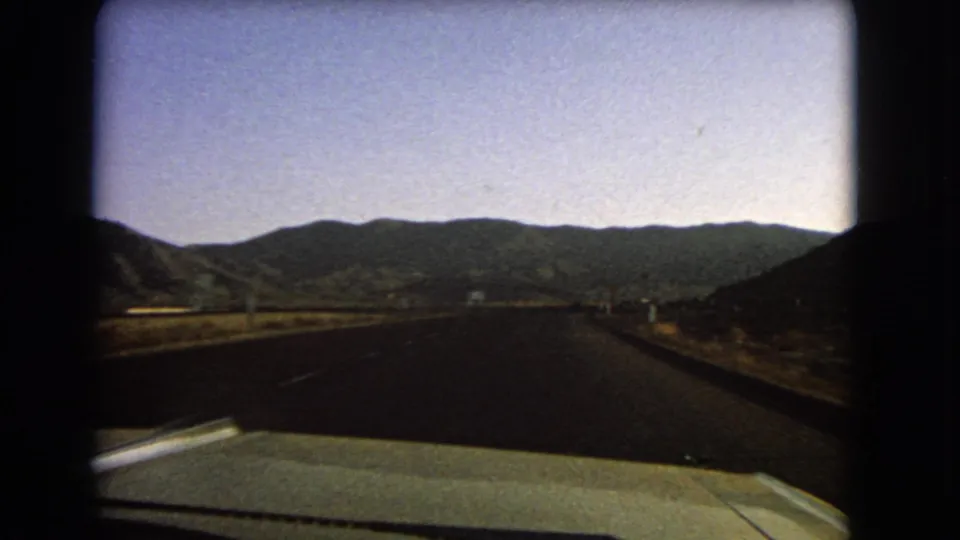 In-car 8mm view of a Nevada highway continuing toward mountain ridges under a pale sky in 1966.