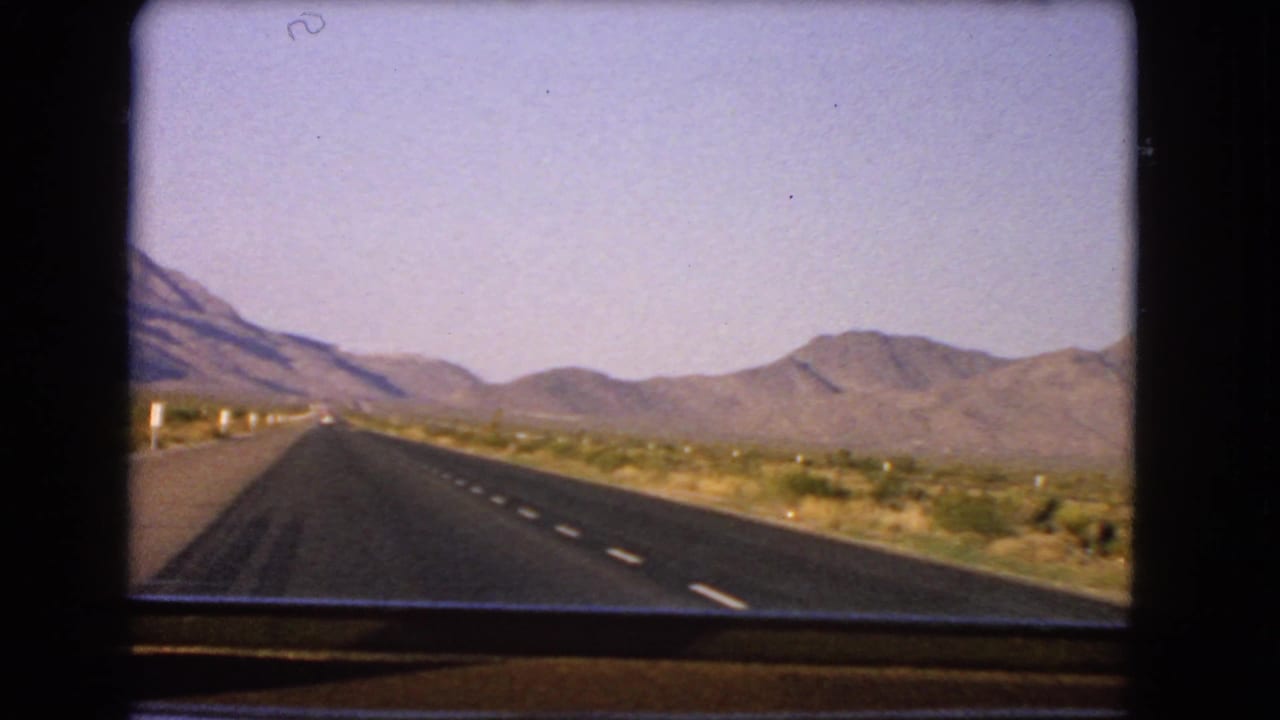 Bright 1966 Nevada desert highway frame showing lane markings, sparse vegetation, and mountains from an in-car perspective.