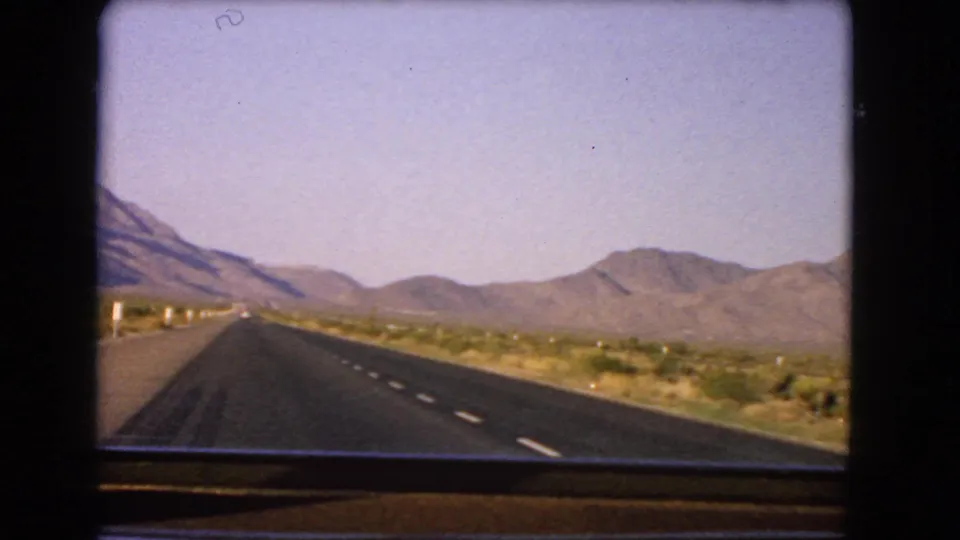 Bright 1966 Nevada desert highway frame showing lane markings, sparse vegetation, and mountains from an in-car perspective.