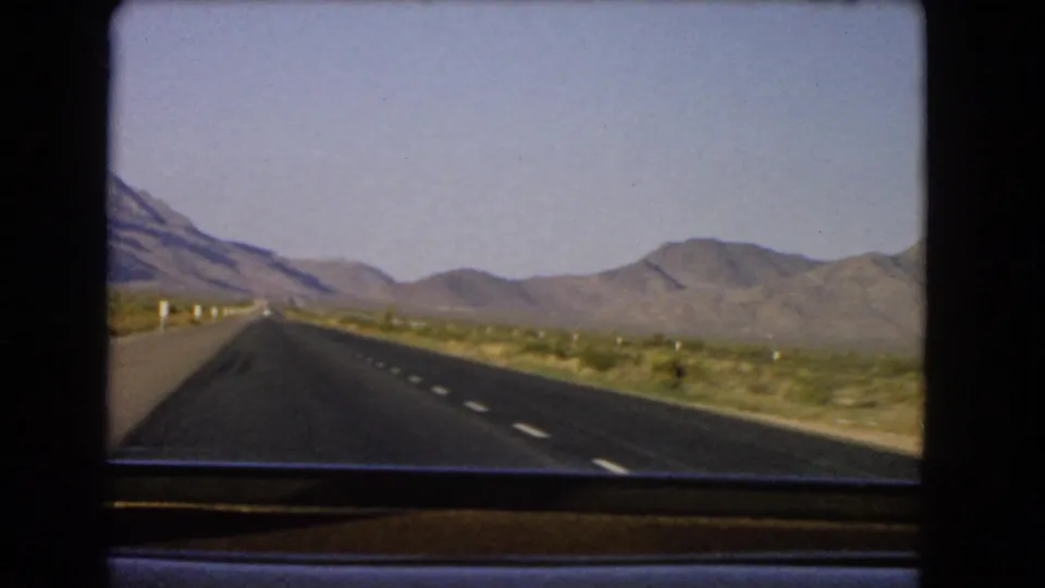 Windshield-view 8mm frame of a Nevada desert highway with scrubland and mountain ridges in the distance.