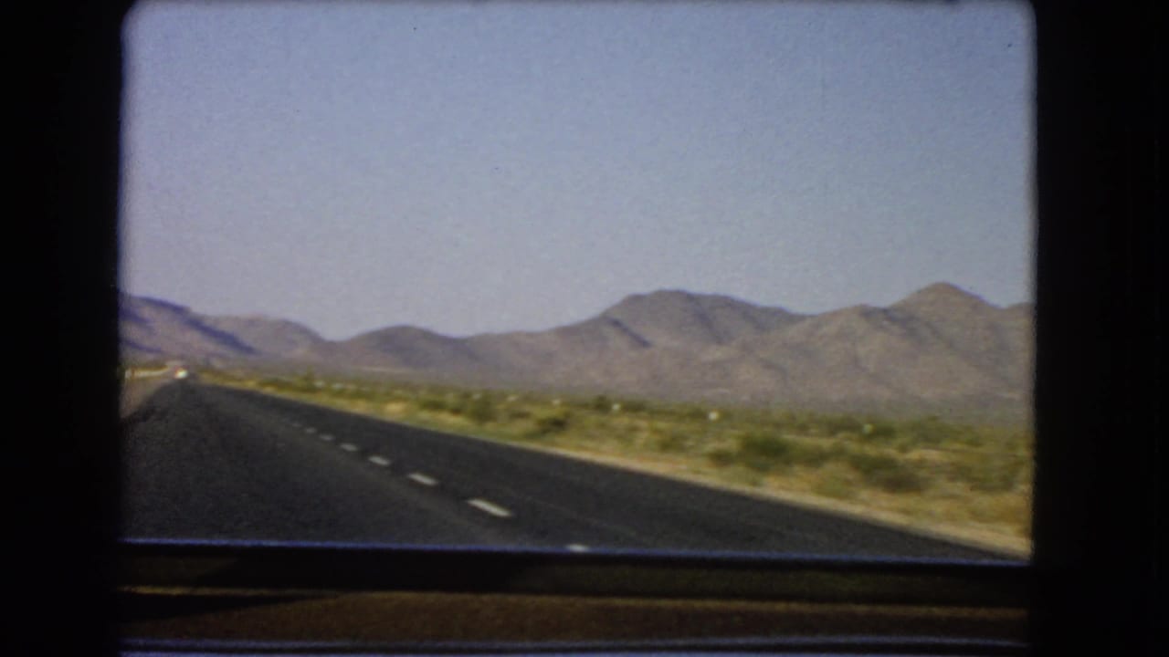 1966 Nevada road-trip frame with the highway shifting left and desert brush filling the roadside foreground.