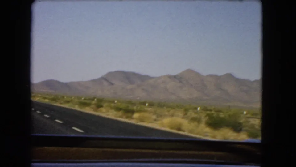 1966 Nevada desert road viewed from inside a car, with sparse scrub and mountains under a clear sky.