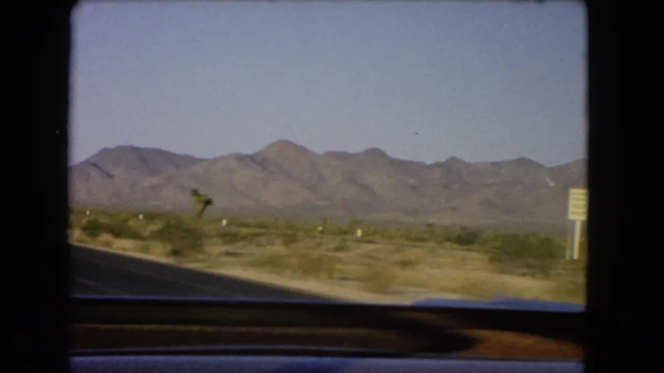 1966 Nevada highway scene with desert scrub, distant mountains, and a yellow roadside sign passing at frame right.