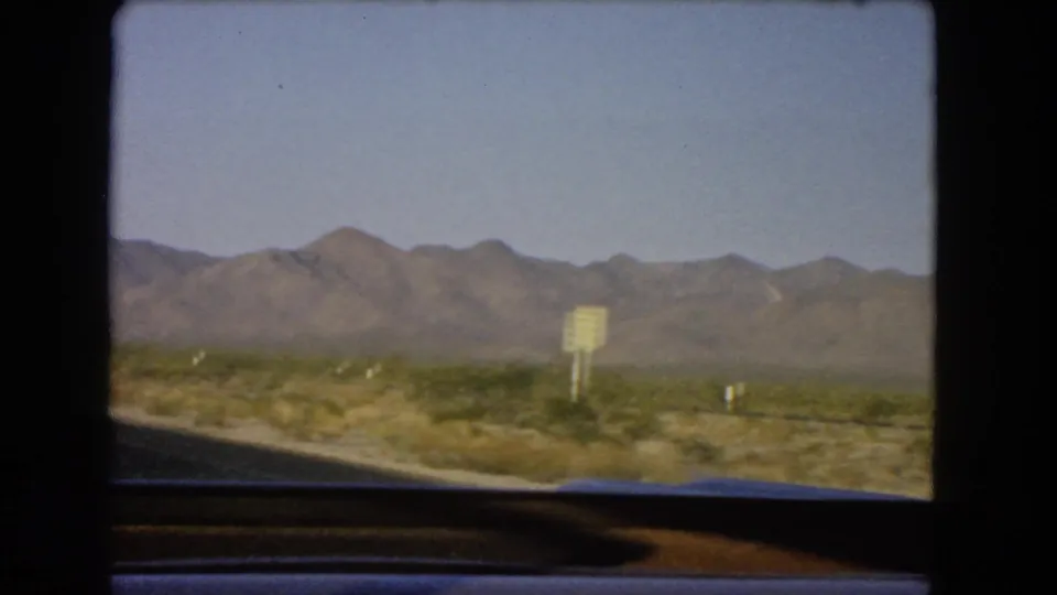 Nevada desert landscape from a moving car in 1966, showing mountains, scrub, and a roadside sign beyond the window frame.
