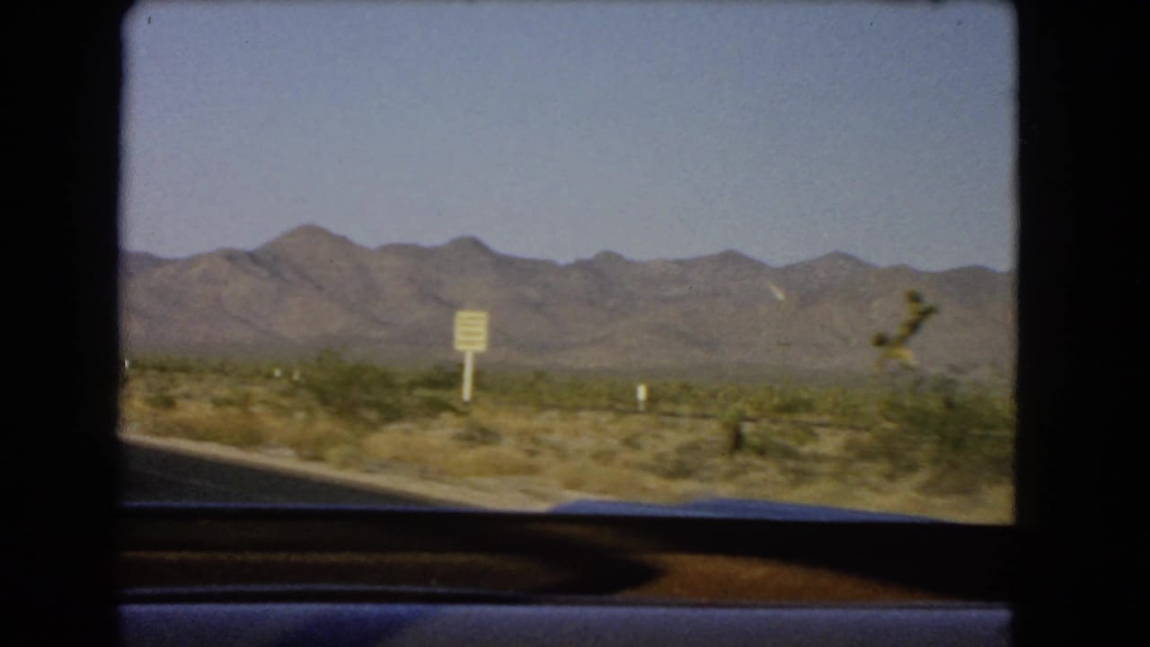 1966 Nevada road-trip view from a moving vehicle showing desert scrub, a roadside sign, and a distant mountain range.