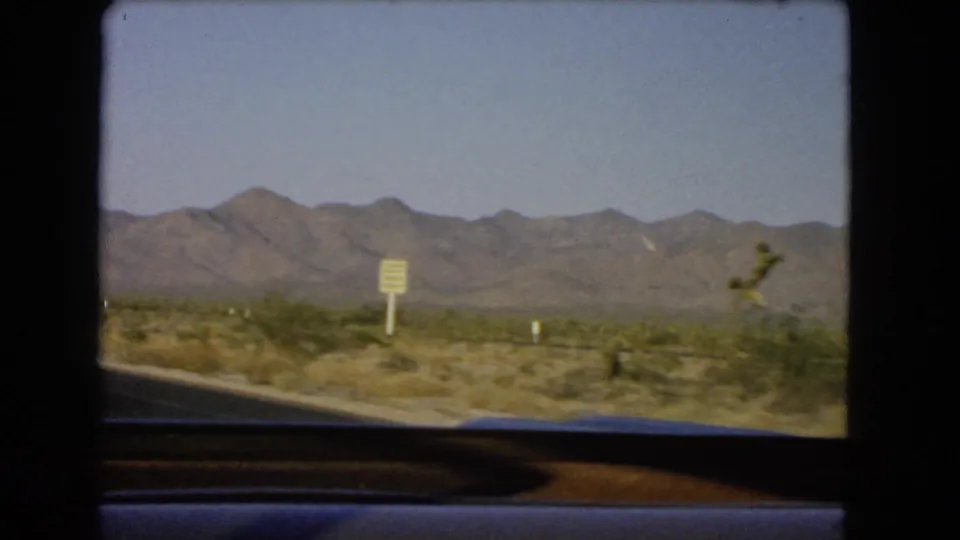 1966 Nevada road-trip view from a moving vehicle showing desert scrub, a roadside sign, and a distant mountain range.