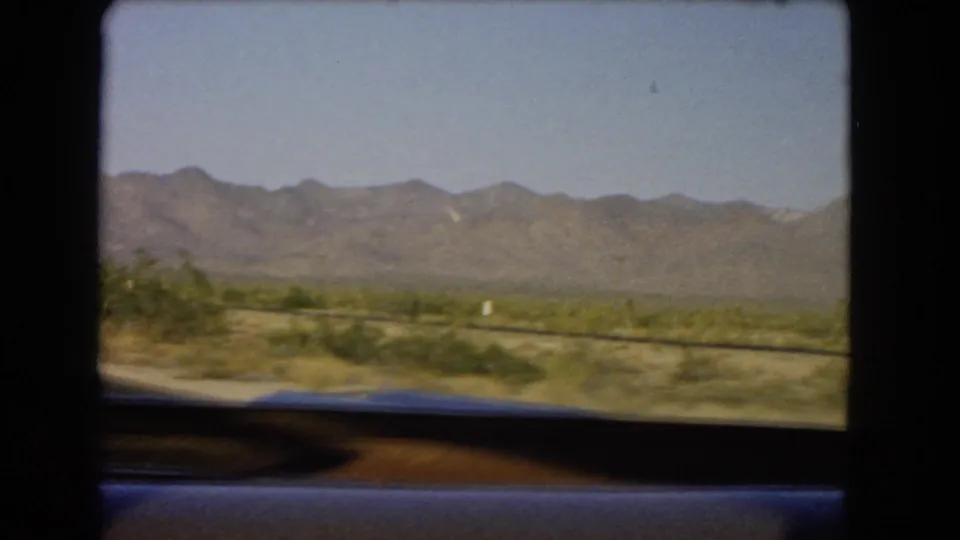 1966 Nevada car-window view across open desert terrain toward a hazy mountain range with road edge in the foreground.