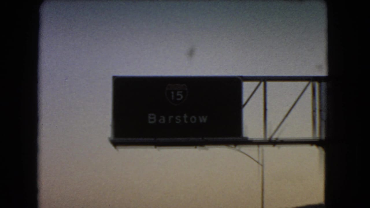 1966 8mm road-trip frame showing an Interstate 15 Barstow sign partly framed by dark vehicle or overpass edges.