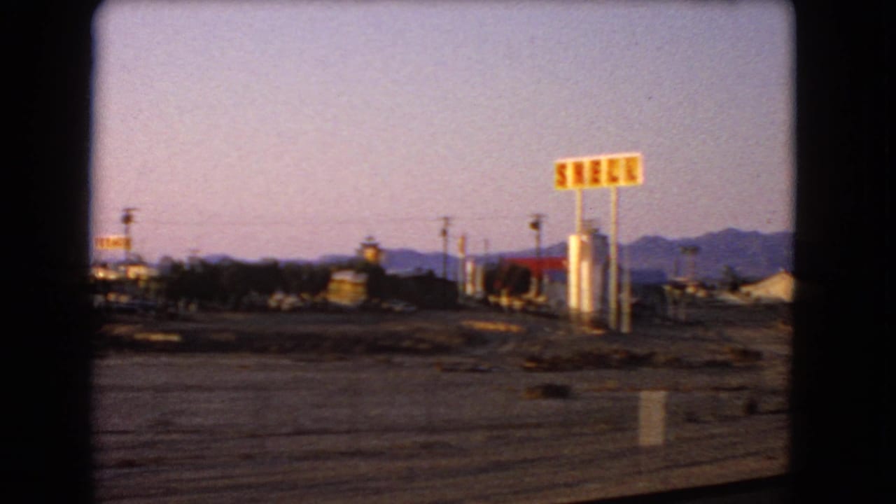 1966 8mm view of a desert roadside with Shell signage, low mid-century buildings, open lots, and mountains in the distance.