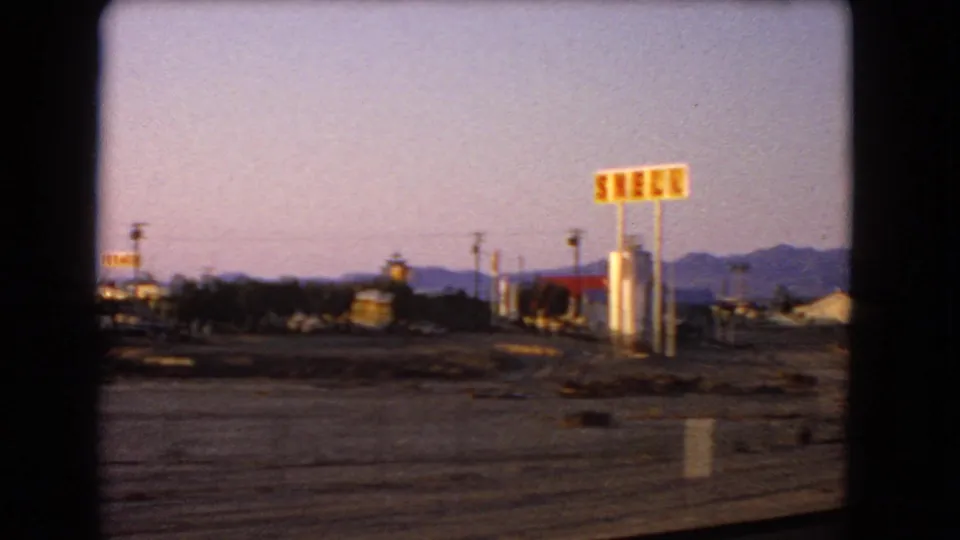 1966 8mm view of a desert roadside with Shell signage, low mid-century buildings, open lots, and mountains in the distance.