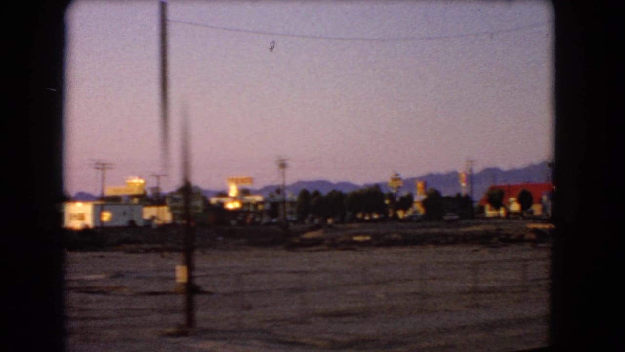 Dusk view from a moving vehicle toward low buildings, lit signs, power poles, and distant mountains.