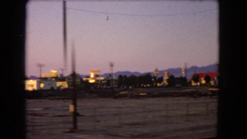 1966 Nevada 8mm frame looking across dusty roadside ground toward a small town edge with illuminated signs and mountains at dusk.