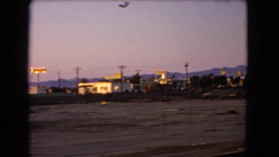 1966 Nevada roadside view with low commercial buildings, bright signs, utility poles, and a mountain horizon at dusk.