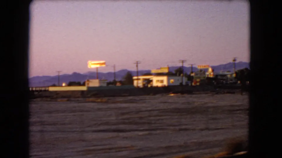 1966 Nevada travel frame showing a passing roadside sign, low town buildings, power poles, and mountains in evening light.