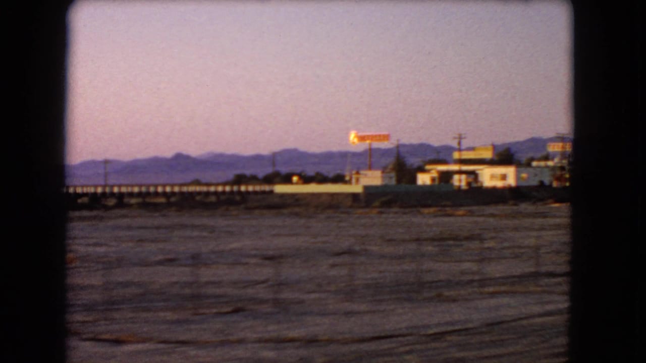 Desert flats, distant mountains, a long bridge, and roadside buildings seen through a dark car window.