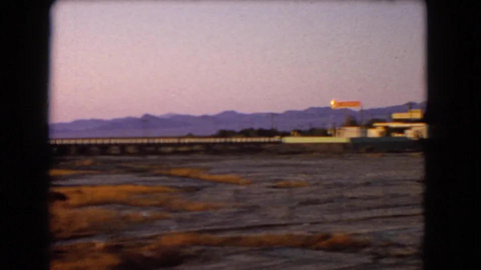 1966 Nevada home-movie frame from a moving vehicle, with pink-purple dusk sky over desert flats, a low bridge, distant mountains, roadside buildings, and an illuminated sign.