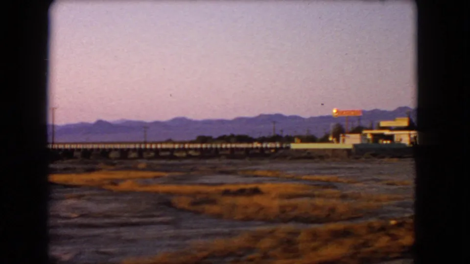 1966 Nevada 8mm frame showing dry scrubby desert foreground, a long bridge or trestle, distant mountain ridges, utility poles, and roadside buildings from a moving car.