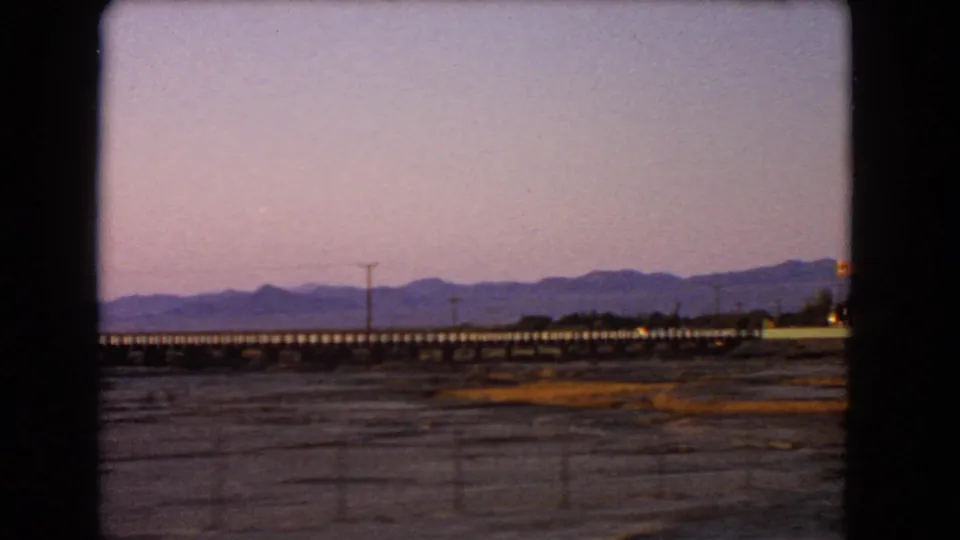 1966 Nevada travel frame with desert flats, a long low bridge, utility poles, distant mountains, and the edge of a roadside building at the far right.