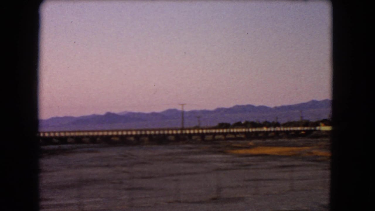1966 Nevada 8mm frame showing a long railway bridge, dry desert terrain, distant mountains, and a purple dusk sky from a moving vehicle.