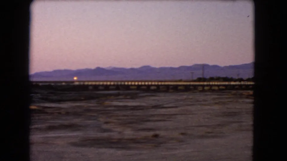 Nevada desert railway bridge at dusk, with a small glowing train light, open dry ground, and mountains under a pink-purple sky.