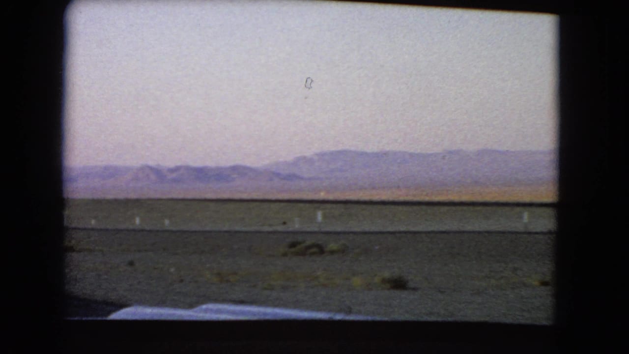 1966 Nevada desert view from a moving vehicle, showing flat roadside terrain, distant mountains, and a low horizontal rail or road line.