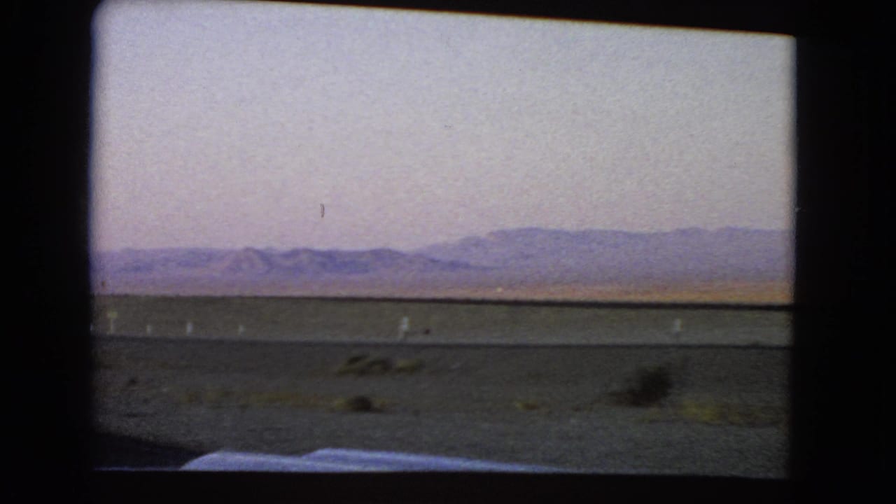 1966 Nevada desert landscape with sparse shrubs and distant hills, filmed through a moving vehicle window on 8mm.