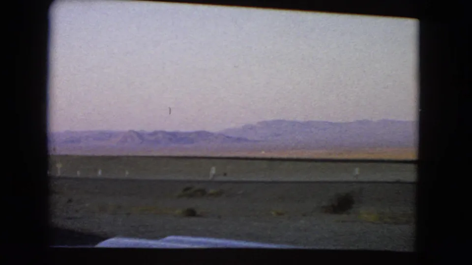 1966 Nevada desert landscape with sparse shrubs and distant hills, filmed through a moving vehicle window on 8mm.