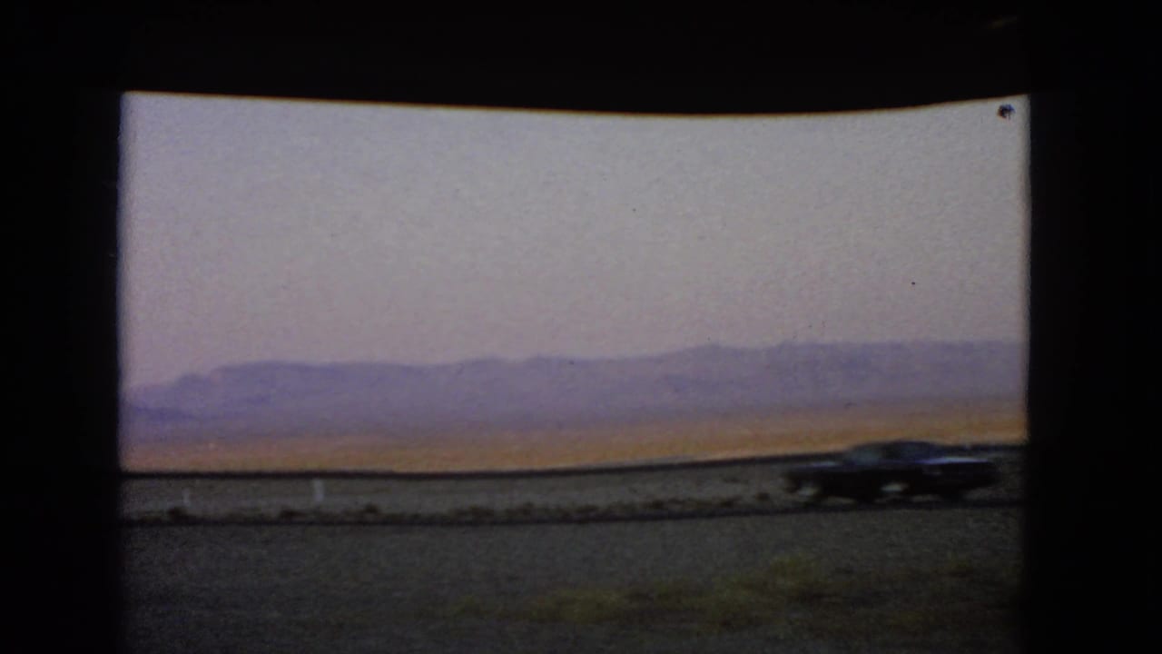 A dark car moves across the Nevada roadside view, with desert flats and mountains behind it.