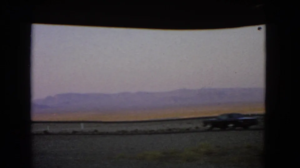 A dark car moves across the Nevada roadside view, with desert flats and mountains behind it.