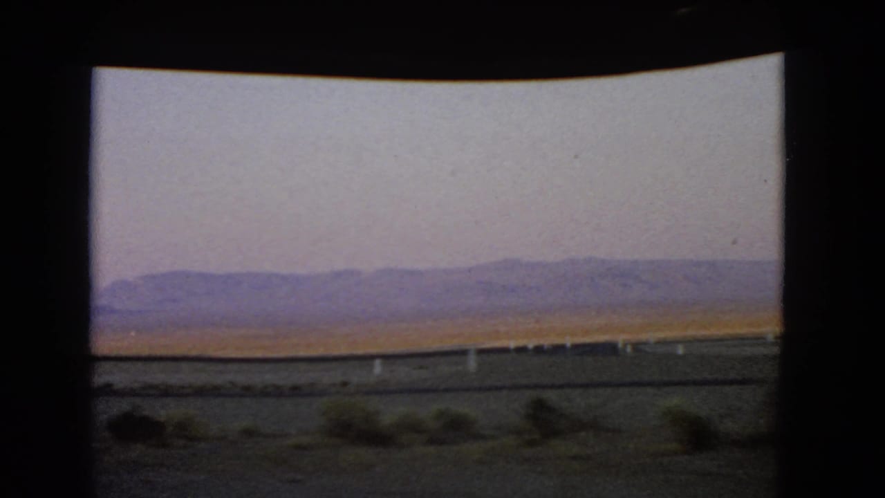 1966 Nevada desert landscape viewed through a moving car window, with roadside posts and hazy mountains beyond the plain.
