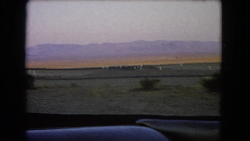 Passenger-side view from a 1966 Nevada car journey, showing open desert, sparse shrubs, roadside markers, and a purple mountain horizon.